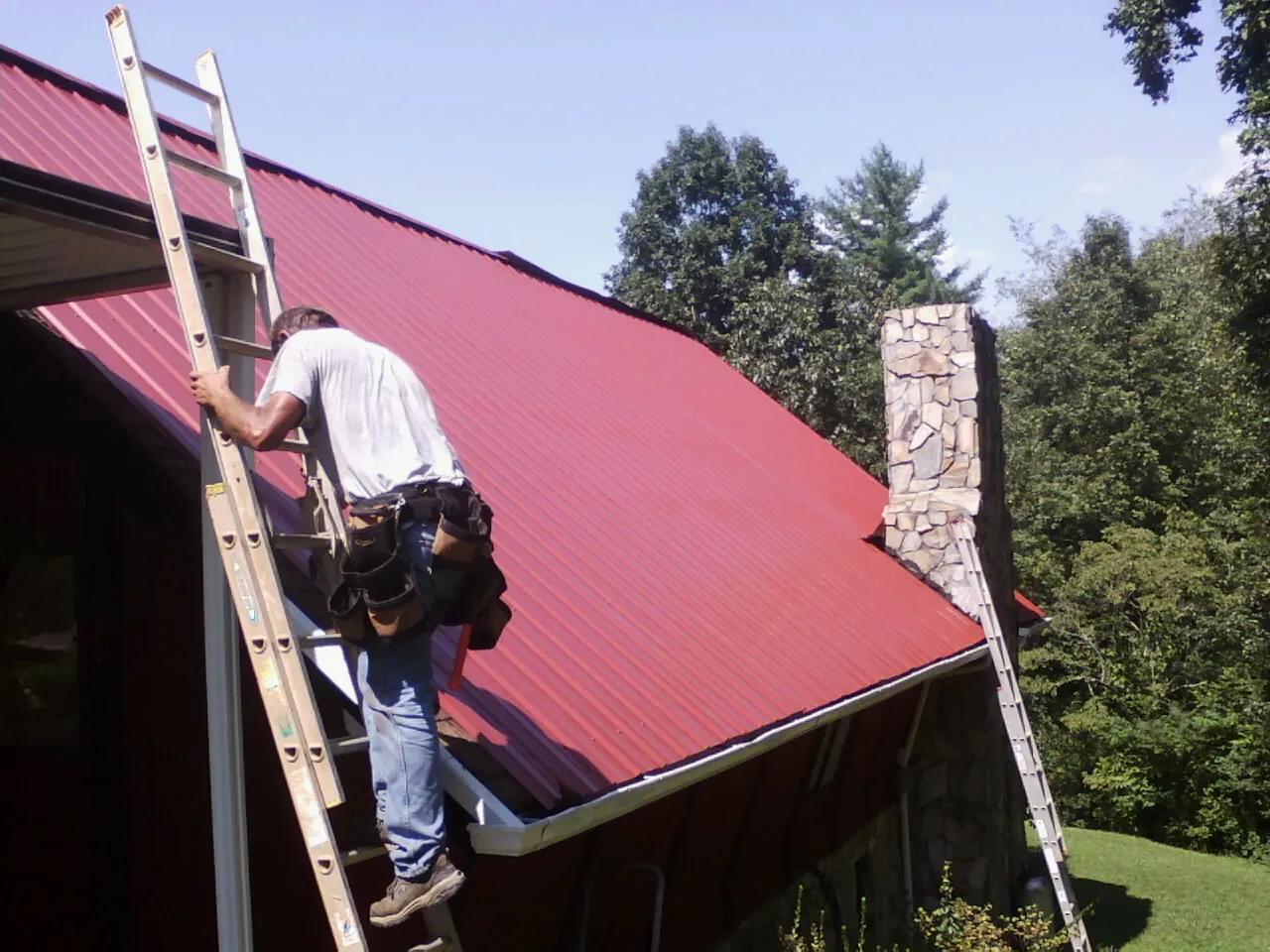 Roofer on a ladder beside a red metal roof and stone chimney — Soffit & Fascia Repair by Roofing Brothers in West Shokan